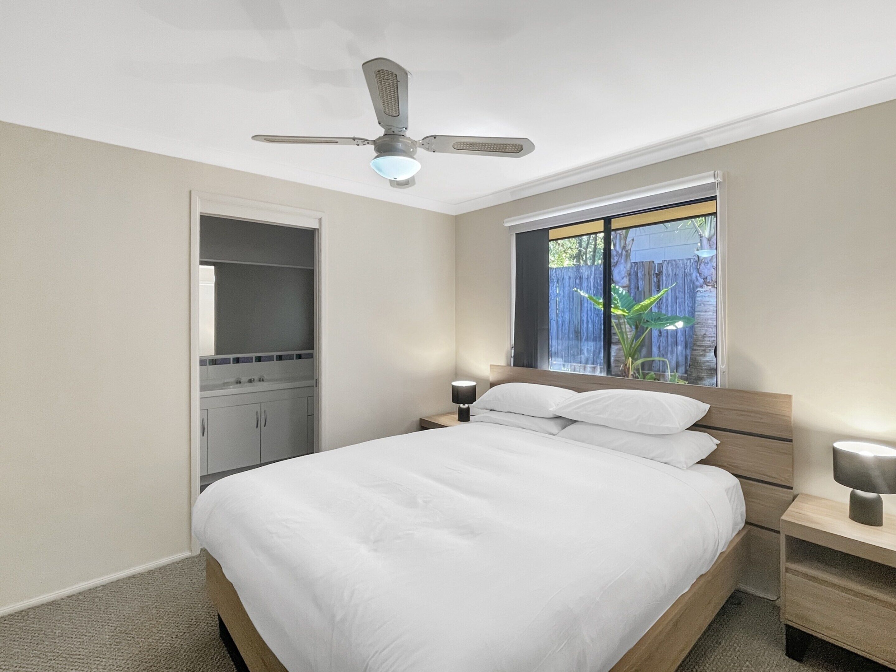 A bedroom with a bed, bedside tables, a ceiling fan, a window with a view of a wooden fence, and a closet.