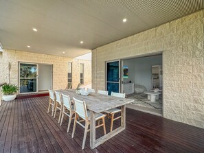 Uma área de refeições coberta ao ar livre com uma mesa e cadeiras de madeira, um vaso de planta e vista do interior através de uma porta de vidro.