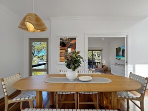 Uma área de refeições com mesa de madeira, cadeiras brancas, um vaso grande com plantas e um lustre.