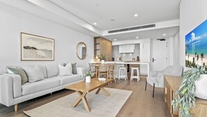 Un salon moderne comprenant un canapé, une table basse et un coin repas. La cuisine est dotée de meubles blancs et d’une grande fenêtre.