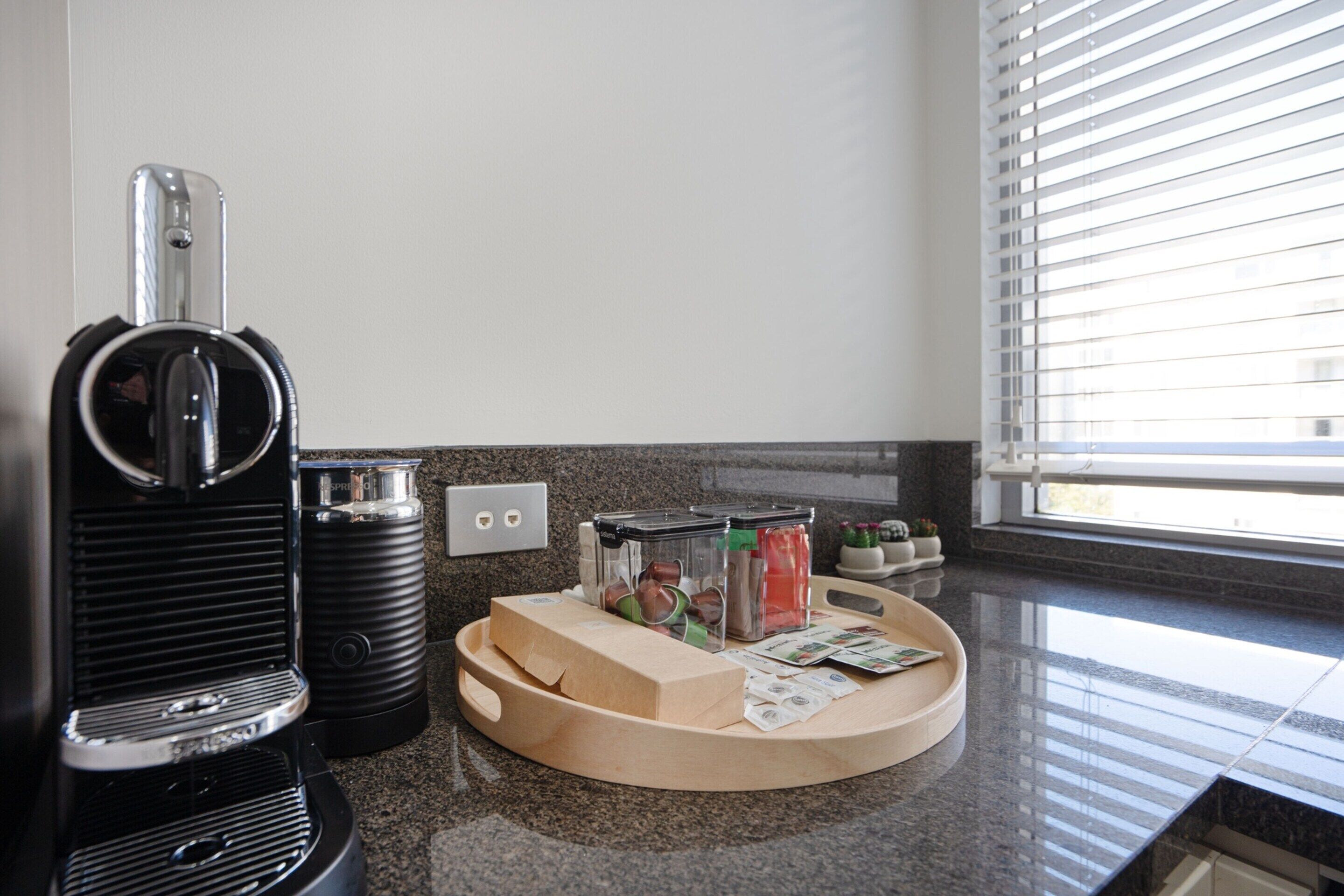 A kitchen area with a coffee maker, a fruit basket, and a window with blinds.