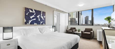 A modern hotel room with a large bed, a desk, a chair, a small table, and a large window offering a city view.