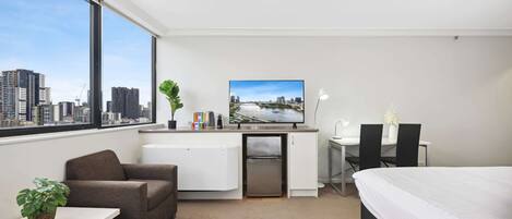 A hotel room with a bed, a desk with a computer, a chair, a small fridge, a TV, and a large window with a city view.