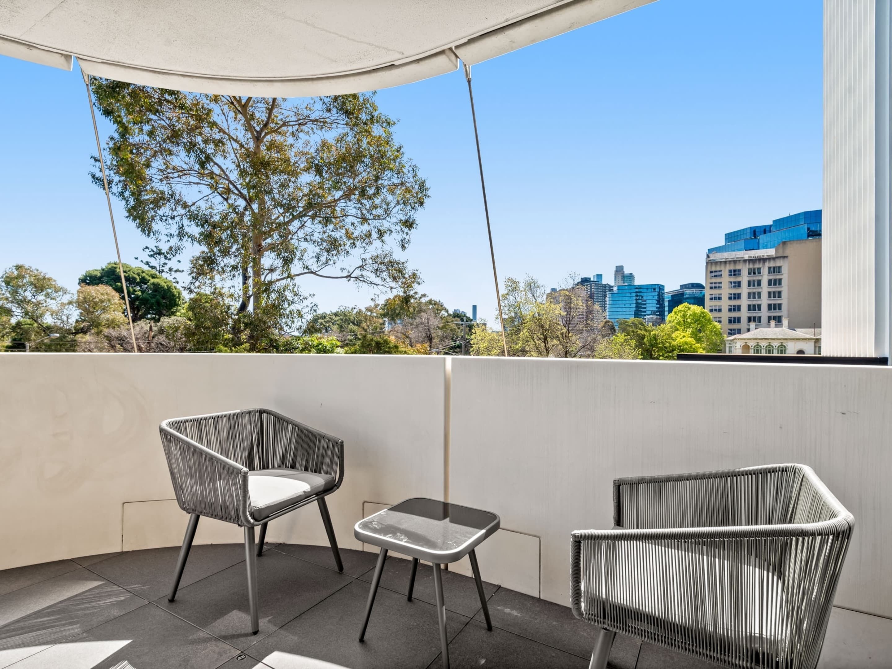 Een balkon met twee rieten stoelen en een kleine tafel, met uitzicht op een stadsgezicht met gebouwen en bomen.