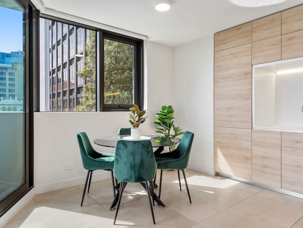 A modern kitchen with a dining area, featuring a round table with chairs, a potted plant, and a large window offering a view of buildings and trees. - Light Filled Apartment by Albert Park Lake (Melbourne)