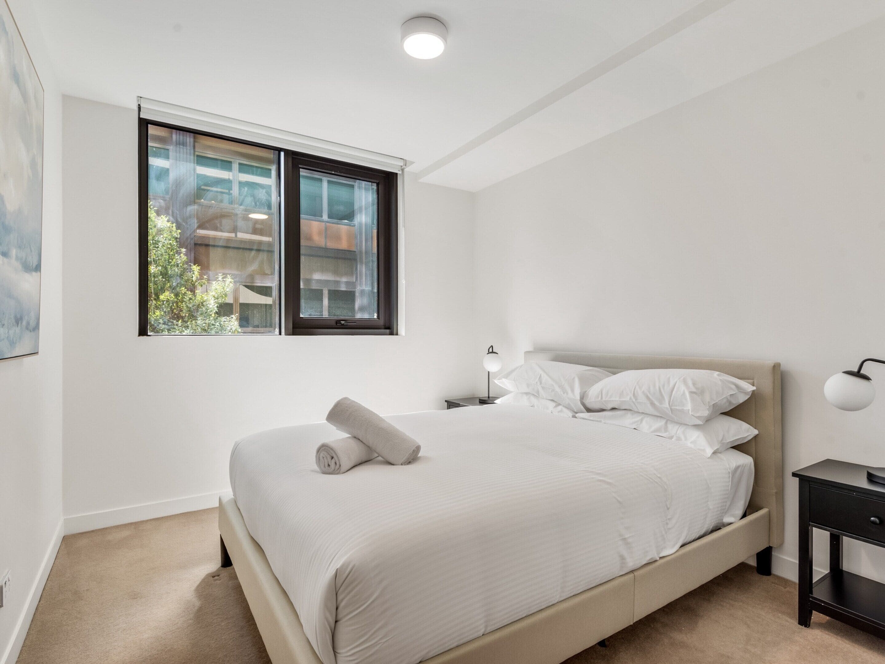 Un dormitorio con cama, mesita de noche, lámpara y ventana con vistas a edificios y árboles.