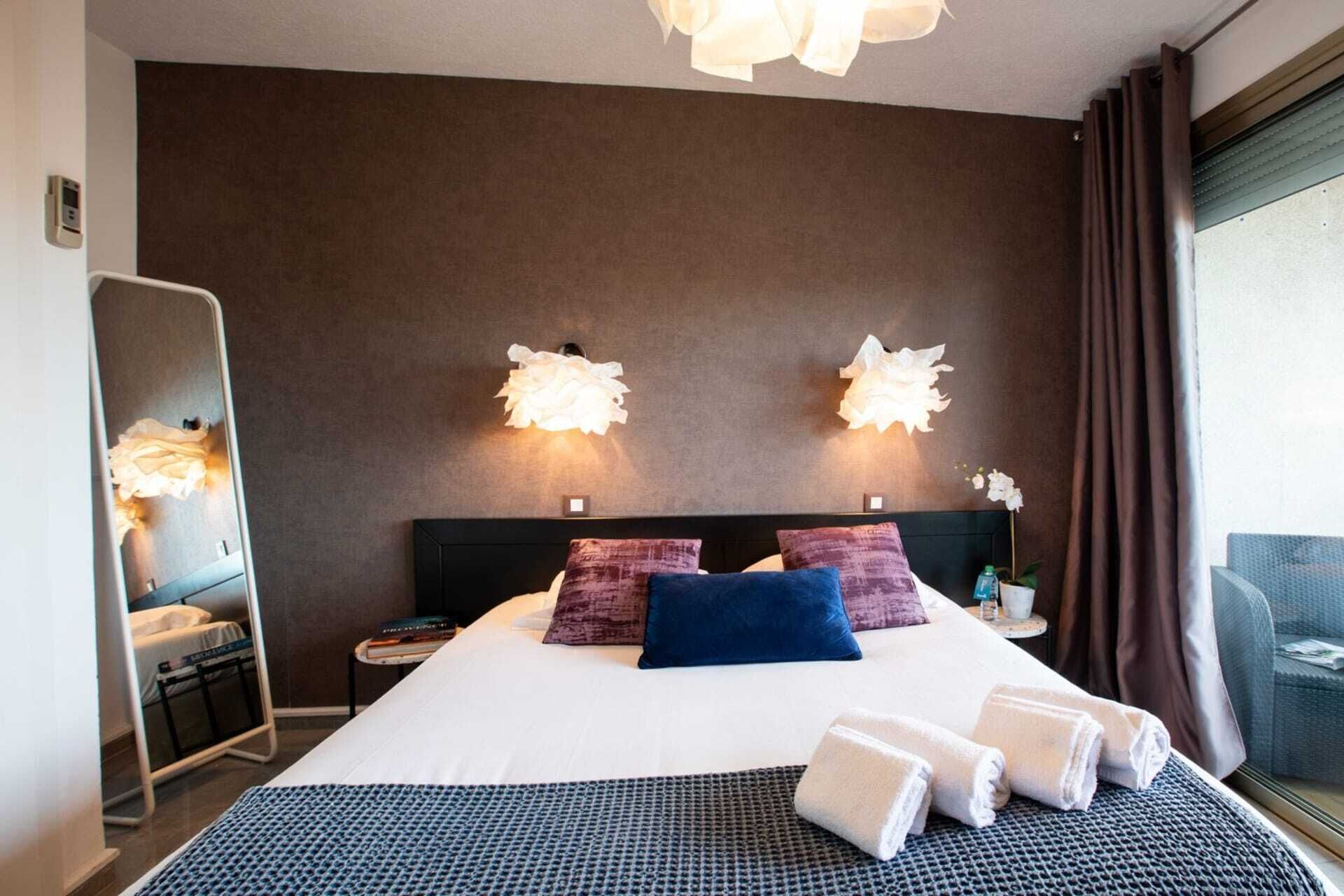 A hotel room with a large bed, two wall-mounted lamps, a nightstand, a mirror, and a window with curtains.