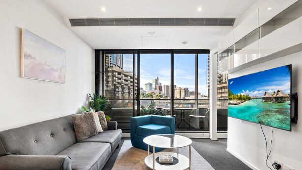 A modern living room with a grey sofa, a blue armchair, and a white coffee table. A large TV is mounted on the wall, and there is a view of a cityscape through large windows.
