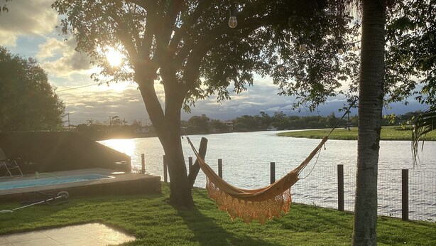 Un hamac suspendu sous un arbre, près d’une piscine, avec vue sur un lac et des bâtiments éloignés.