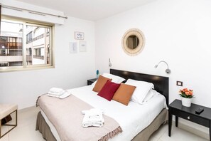 A hotel room with a bed, a bedside table with a lamp, a small table with a vase of flowers, and a window with a view of buildings.