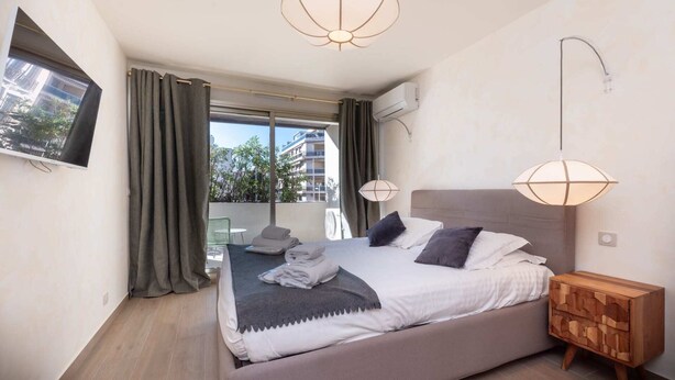 A modern bedroom with a bed, bedside lamps, a wooden nightstand, and a view through the window. - Two-bedroom 1B - Two Bedroom Apartment, Sleeps 6 (Antibes)