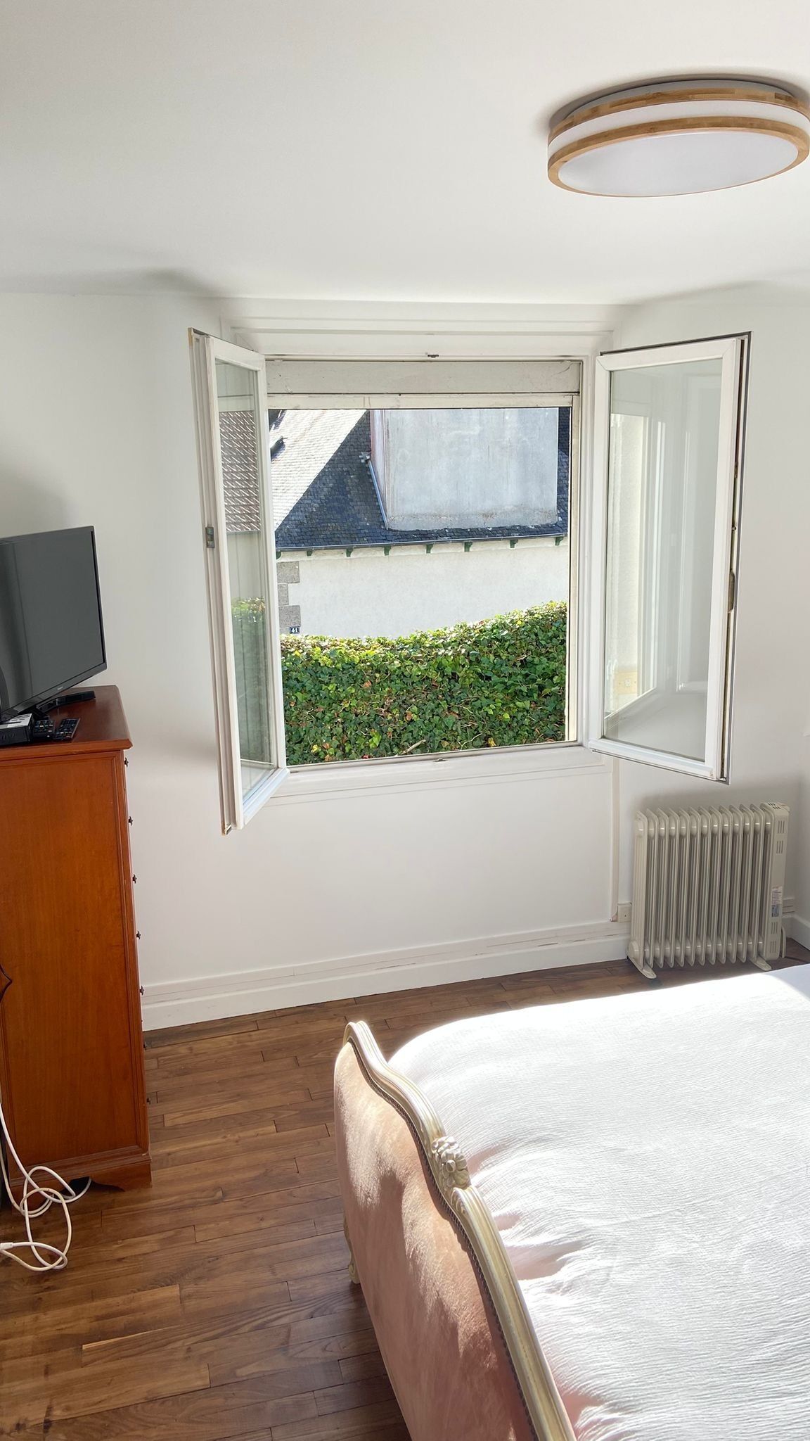Et soveværelse med en stor seng, et fjernsyn på et træskab, en radiator og et vindue med åben persienne.