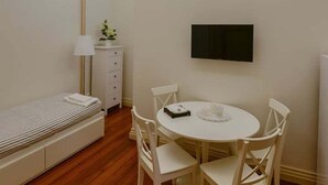 Ein kleiner, hell erleuchteter Raum mit einem runden Tisch und vier Stühlen, einem Bett mit gestreifter Decke, einem Nachttisch mit Lampe sowie einem an der Wand befestigten Fernseher.