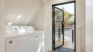 A laundry room with a washer and dryer, a door leading to an outdoor area with a gravel path and trees. - Modern and Secluded A-Frame with a Hot Tub for a Romantic Texas Hill Country Getaway (Wimberley)