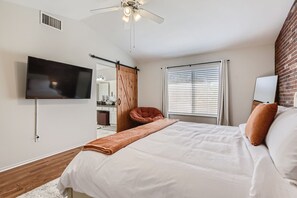 A bedroom with a bed, a TV mounted on the wall, a ceiling fan, a window with blinds, and a brick wall.