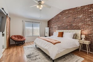 Une chambre avec un mur en briques, un lit, une chaise, un ventilateur de plafond et une fenĂȘtre avec des stores.