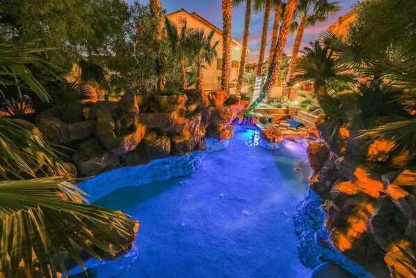 A pool with a blue hue, surrounded by rocks and palm trees, with a building in the background.