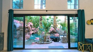 Ein Zimmer mit einer großen Schiebeglasdoor, die zu einem Felsengarten mit Wasserspiel, Sitzgelegenheit und Blick auf Palmen führt.