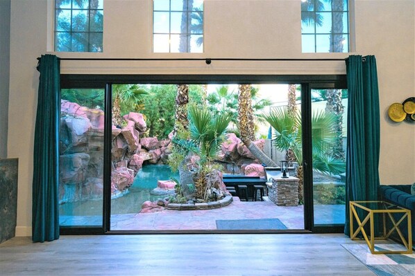 A room with a large sliding glass door leading to a rock garden with a water feature, seating area, and a view of palm trees. - OMG! Wild Oasis! Amazing Pool! Modern Villa! (Spring Valley)