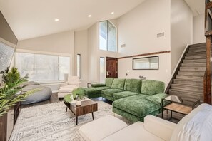Un salón amplio con un sofá seccional verde, un sillón blanco, una mesa de centro y una escalera.