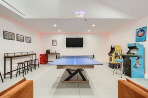 A room with a ping pong table, bar stools, a TV, arcade machines, and framed pictures on the wall.