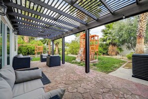 An outdoor patio with stone flooring, a covered seating area with sofas, and a playground structure.