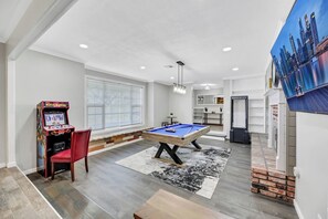 A pool table in a well-lit room with a window, a red chair, and a video game machine.