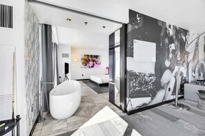 Ein modernes Hotelzimmer mit einer großen Badewanne, einem Bett, einem an der Wand montierten Fernseher und einem großen abstrakten Gemälde.