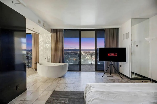 A modern hotel room with a large bathtub, a TV, and a balcony with a city view.
