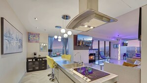 Una cocina moderna con una isla central, una zona de comedor con sillas amarillas, una chimenea y vistas a la ciudad.