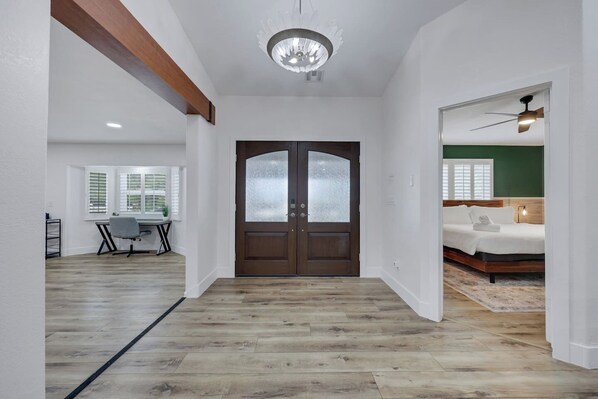 A hallway leading to a bedroom with a bed, a desk with a chair, and a window with blinds.