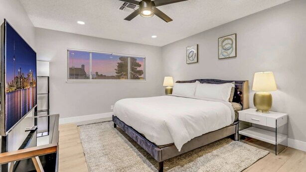Um quarto com uma cama, duas mesas de cabeceira com luminárias, um ventilador de teto, uma TV em um móvel e obras de arte na parede.