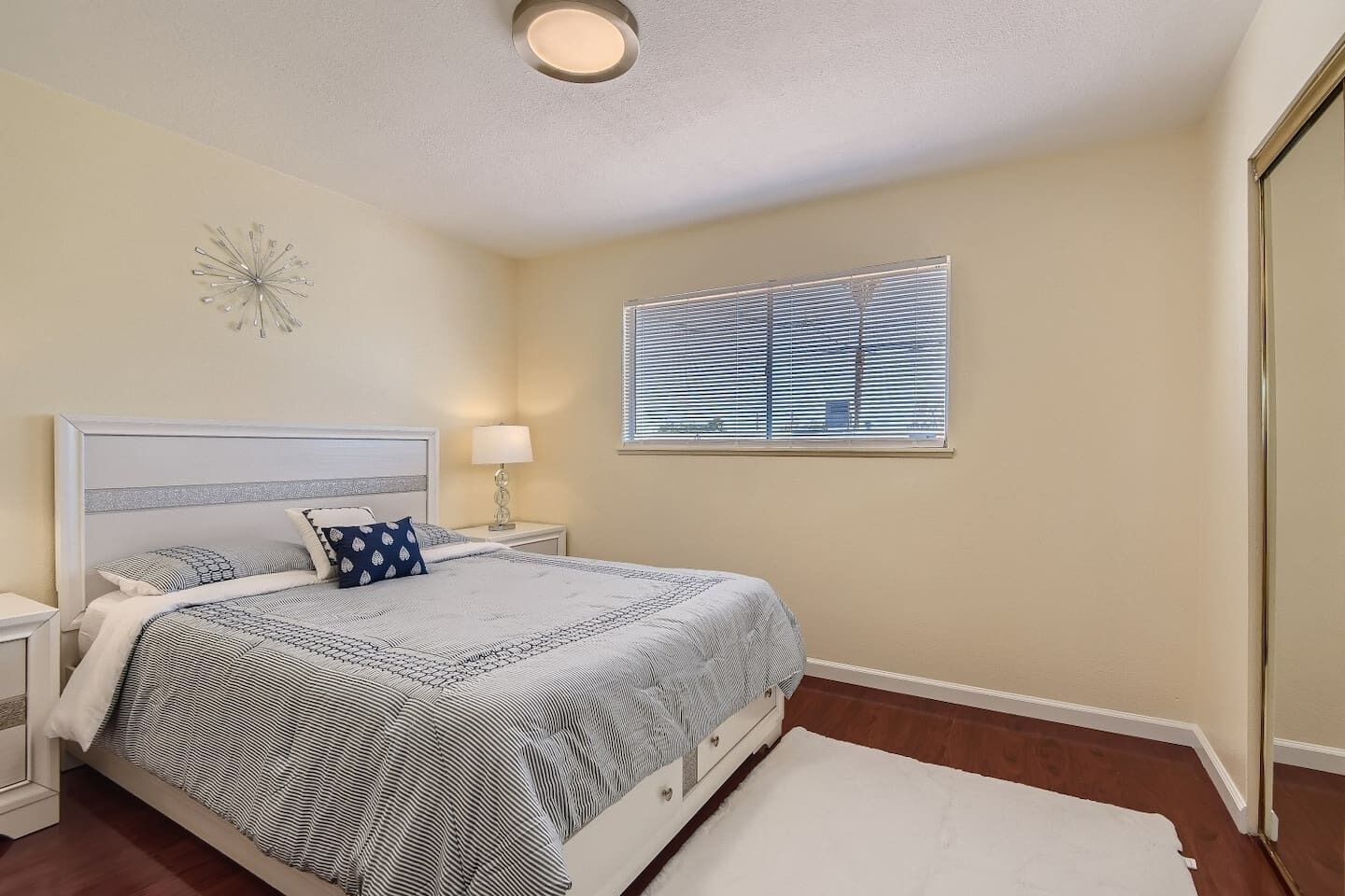 A bedroom with a bed, nightstands, a lamp, a window with blinds, and a ceiling light.