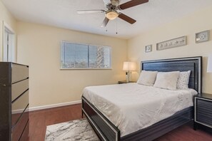 Une chambre à coucher comprenant un lit, un ventilateur de plafond, une table de chevet avec une lampe, une commode et une fenêtre avec des stores.