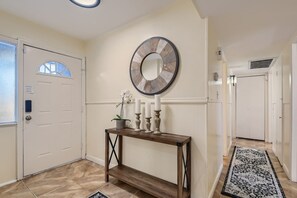 Un couloir avec une porte blanche, une console ornée de bougies, une horloge murale et un tapis à motifs.