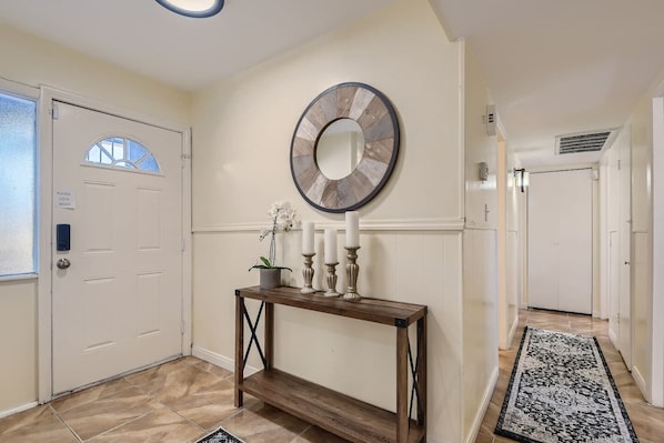 Un couloir avec une porte blanche, une console ornée de bougies, une horloge murale et un tapis à motifs.