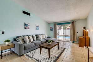 A living room with a grey sofa, a wooden coffee table, a small side table with a plant, and a view of a swimming pool through a glass door.