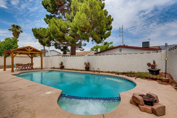 A swimming pool with a wooden pergola and chairs, surrounded by a white wall and trees.