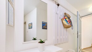A white bathroom with a large mirror, a potted plant, and a wall-mounted basket with colorful items.