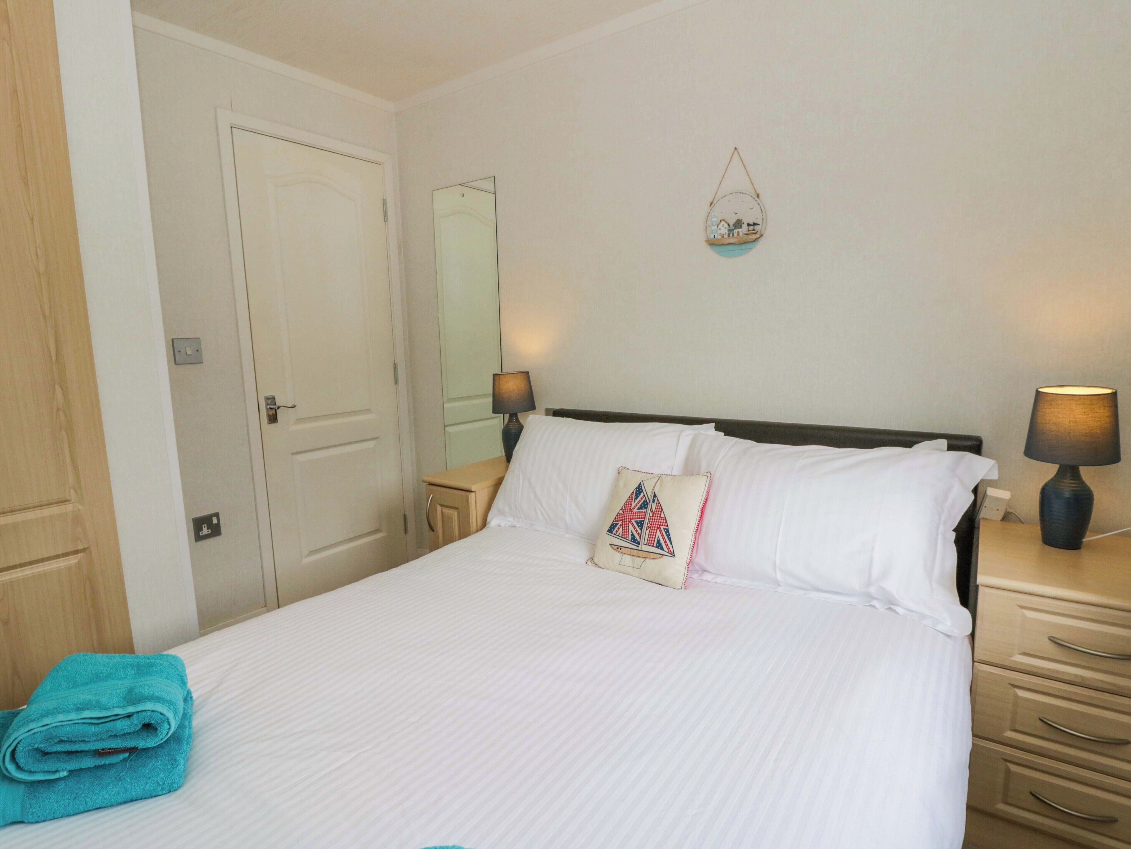 Een net opgemaakt bed met witte lakens, een opgevouwen blauwe handdoek, een bedlamp en een spiegel aan de muur.