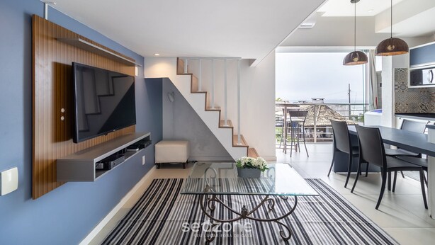A modern living room with a flat-screen TV, a staircase, a dining area, and a view of the city.
