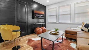 A modern living room with a black wall, a TV displaying "WELCOME TO CHARLOTTE," a yellow chair, a brown sofa, and a wooden floor.