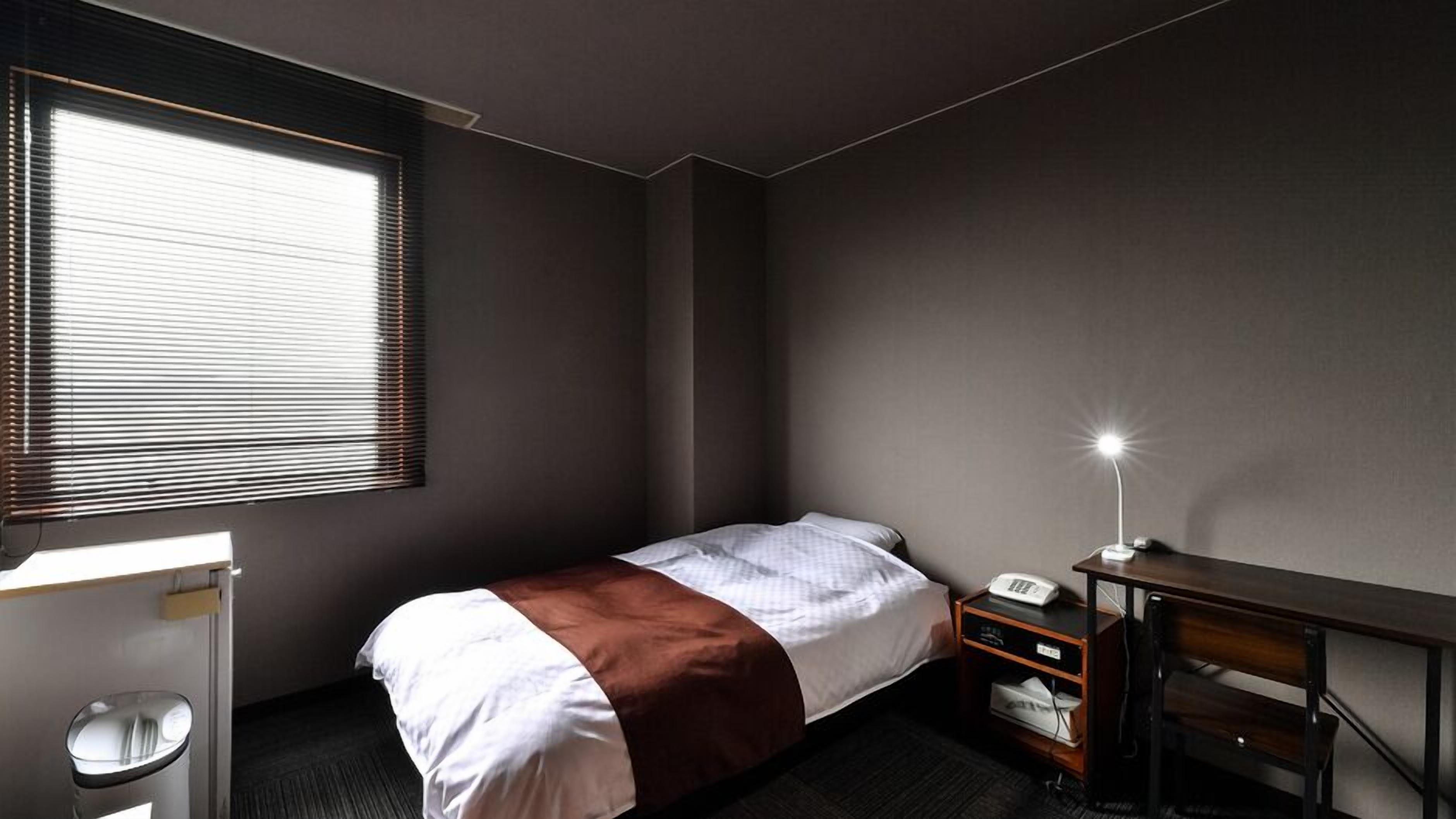 A single bed with a white and brown blanket, a desk with a lamp, a telephone, and a trash bin in a hotel room.