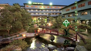 A traditional Japanese garden with a bridge, koi pond, and buildings in the background. - Awara Onsen Mimatsu (Awara)