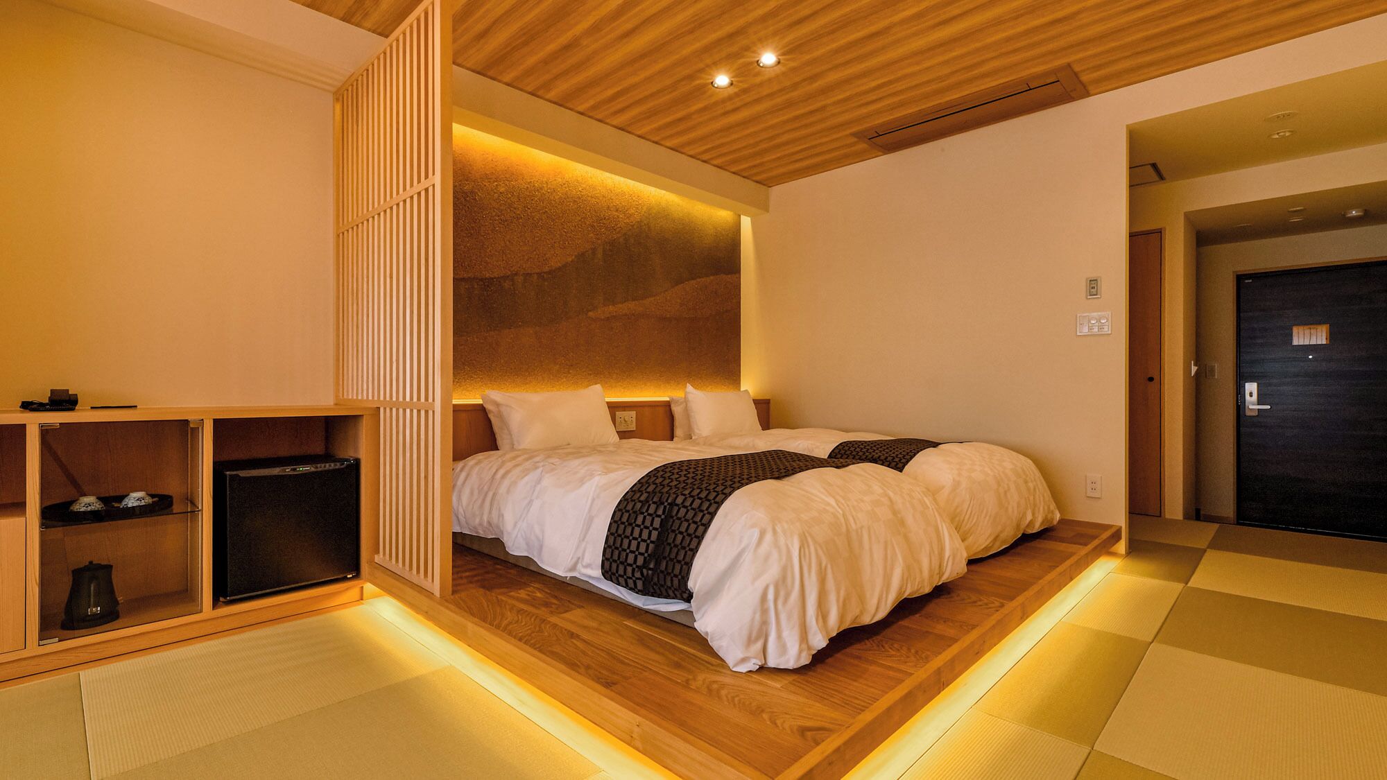 A hotel room with a bed, wooden flooring, a wall-mounted shelf, and a door.