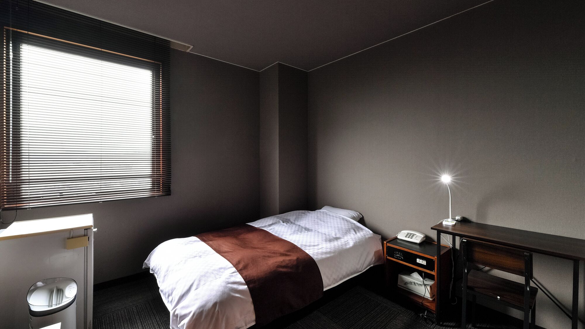 A small, neatly arranged hotel room with a single bed, a desk, a chair, a bedside table, a lamp, and a telephone.
