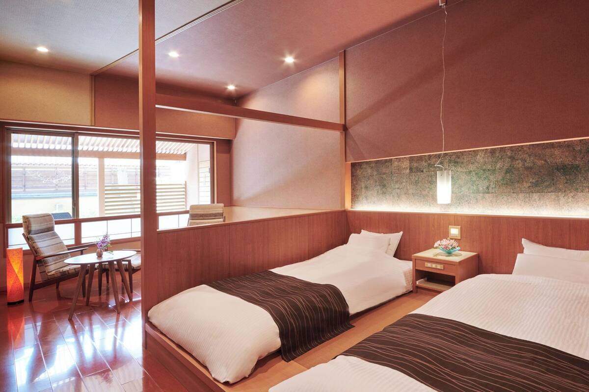 A hotel room with two beds, a small table, a chair, and a large window.