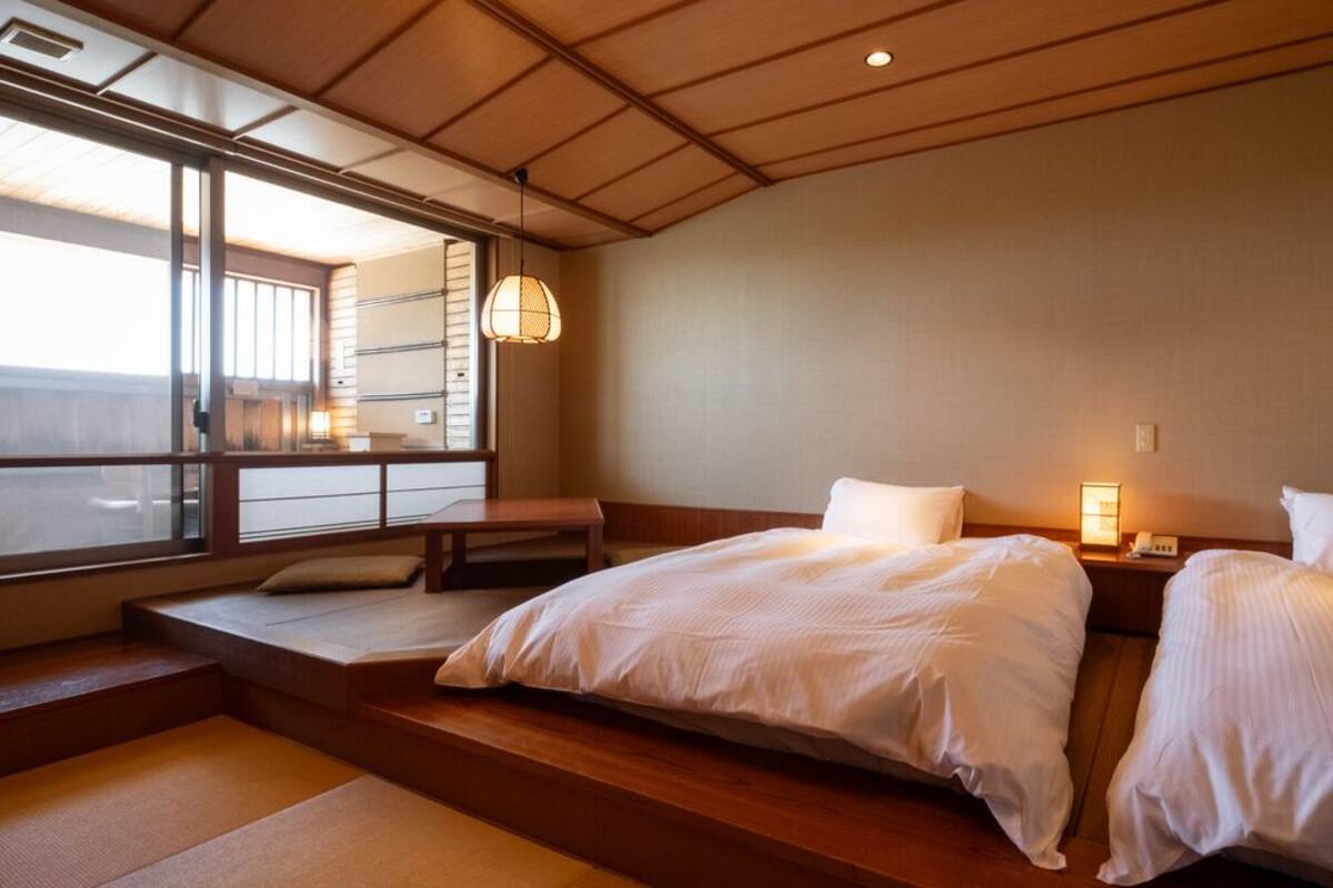 A hotel room with two beds, a bench, a large window, and a hanging light fixture.