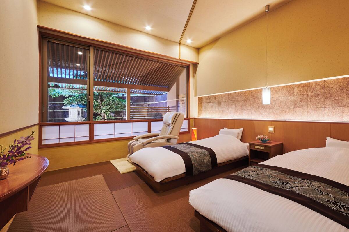 A hotel room with two beds, a sitting area with a chair, a small table, and a view of the outside.