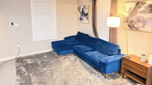 A room with a blue sectional sofa, a wooden nightstand, a lamp, and a ceiling fan. - Cozy 2-bedroom bungalow in cozy East Point GA 15 mins from downtown Atlanta (East Point)